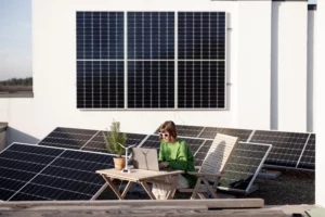 A young woman working on her laptop sits at a small outdoor table outside of her rooftop apartment. Her sides of her apartment and the rest of the roof are covered with solar panels.