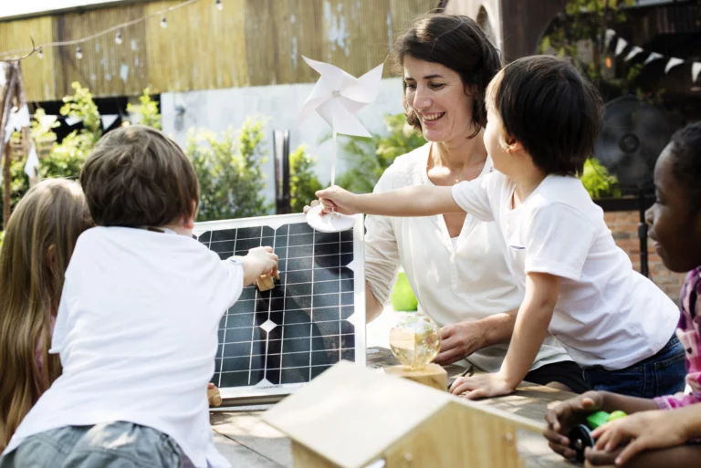 A smiling woman engages a group of children in a hands-on lesson about clean energy involving a solar panel.
